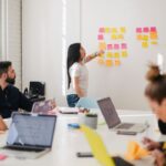 Frau vor Wand mit Post-its im Meeting-Raum mit Kollegen und Kolleginnen.