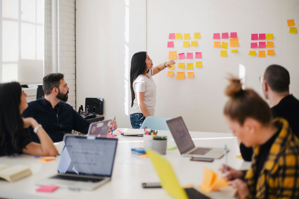 Frau vor Wand mit Post-its im Meeting-Raum mit Kollegen und Kolleginnen.